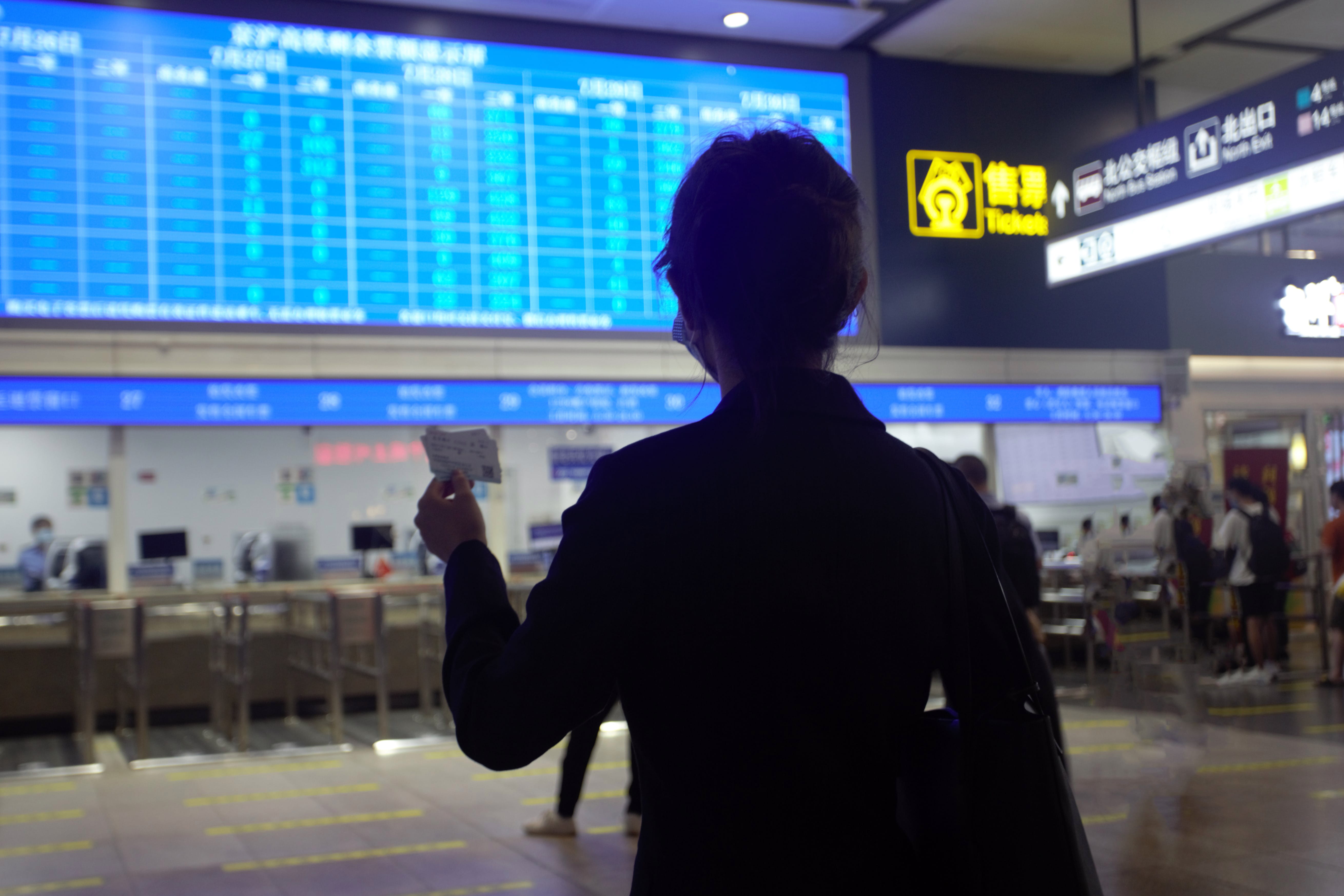 mujer de pie en el aeropuerto frente a la pantalla, buscando el horario de su vuelo con el billete en la mano.