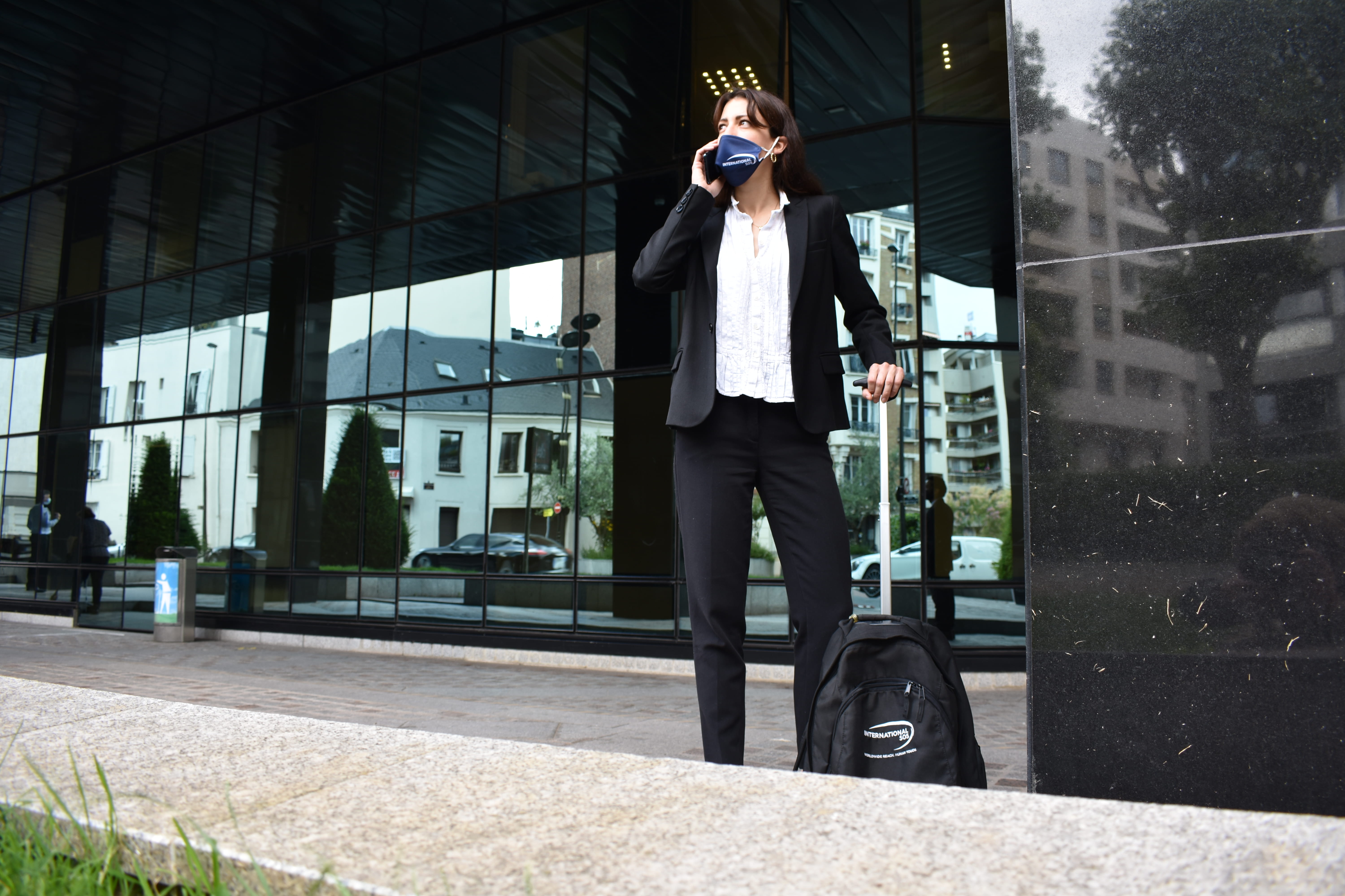 un hombre con máscara y bolsa de viaje se encuentra frente a un edificio usando su teléfono móvil